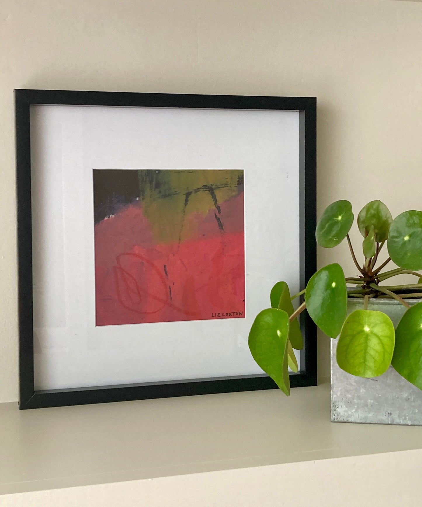 Abstract art by Liz Loxton Studio in square frame on a shelf with a plant. Colours of the painting are pinks and greens.